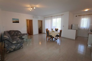Sala de estar con sofá, una mesa de centro de vidrio, una mesa de comedor con sillas, una zona de cocina con refrigerador y una escalera que lleva a otra habitación.