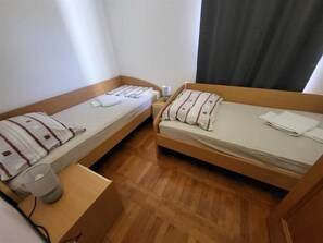 Ett litet, kompakt rum med två enkelsängar, ett trägolv, ett litet bord med en lampa och ett fönster med gardiner.