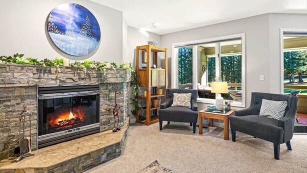 A living room with a stone fireplace, a round wall art piece, a bookshelf, and a large window with a view of trees.