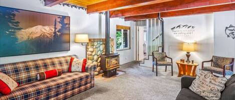 A cozy living room with a plaid sofa, a dark armchair, a stone fireplace, and a painting of a mountain.