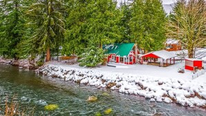 Eine Holzhütte am Fluss, mit schneebedecktem Boden und Bäumen.
