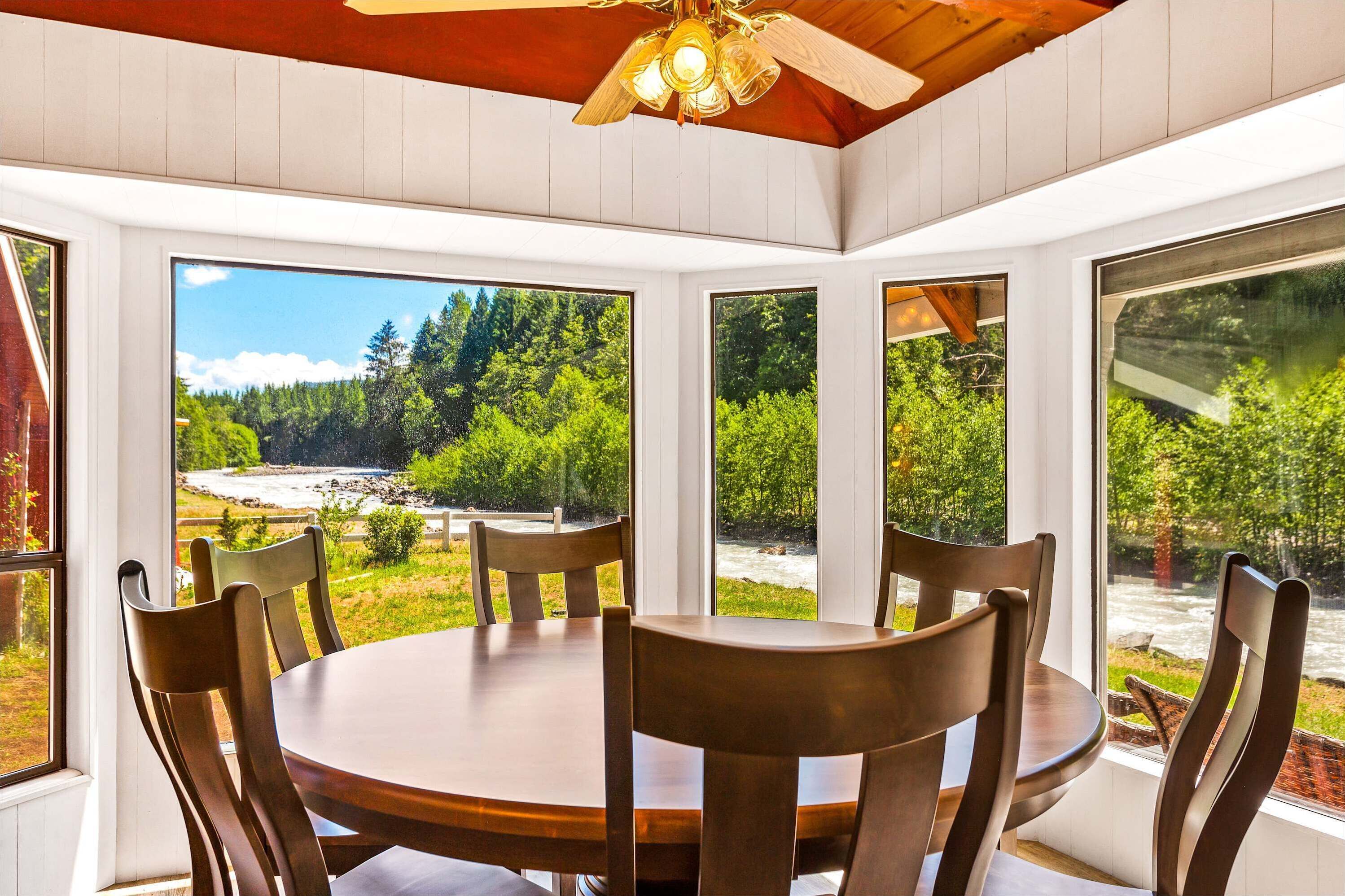 A dining area with a round table and wooden chairs, large windows with a view of a forest and a river, and a ceiling fan.