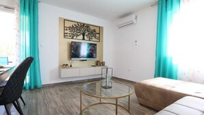 Een moderne woonkamer met een televisie, een ronde glazen salontafel, een beige bank en teal gordijnen.