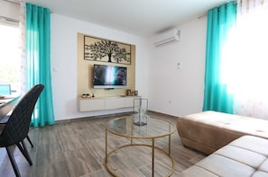 Ein modernes Wohnzimmer mit Fernseher, rundem Glastisch, beiger Couch und blaustichigen Vorhängen.
