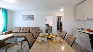 Een moderne woonkamer met een bank, een eettafel gedekt met eten en drinken, een keuken met kastjes en een spoelbak, en een gang die leidt naar een slaapkamer.