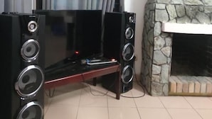 Una sala de estar con televisor de pantalla plana, dos altavoces grandes, chimenea de piedra y una ventana con cortinas.