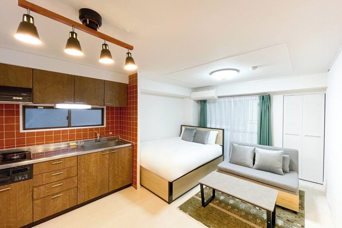 Een compacte hotelkamer met een kitchenette, een bed, een slaapbank en een raam met gordijnen.