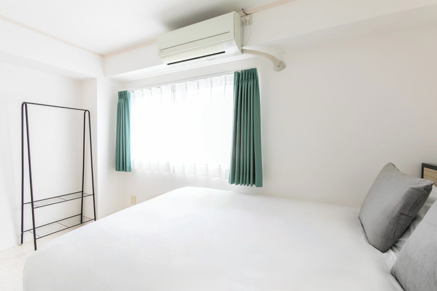 A hotel room with a bed, a window with curtains, an air conditioning unit, and a metal rack.