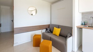 A modern room with a sofa, ottomans, a mirror, a kitchen area, and a door. - L'appartement Cala Bianca (BORMES LES MIMOSAS)