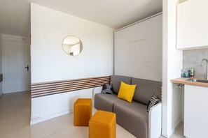 A modern room with a sofa, ottomans, a mirror, a kitchen area, and a door. - L'appartement Cala Bianca (BORMES LES MIMOSAS)