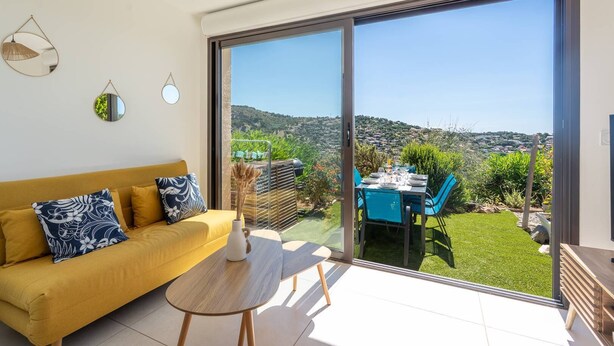 A modern living room with a sofa, a small table, and a view through a sliding glass door to a garden and hillside. - L'appartement Cala Bianca (BORMES LES MIMOSAS)