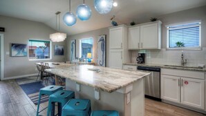 A modern kitchen with a marble island, bar stools, pendant lights, and a view of the outside. - Tickle My Fansea by Avantstay Mins to Shore + Private Pool + Lounge Stylish Beach House (Port Aransas)
