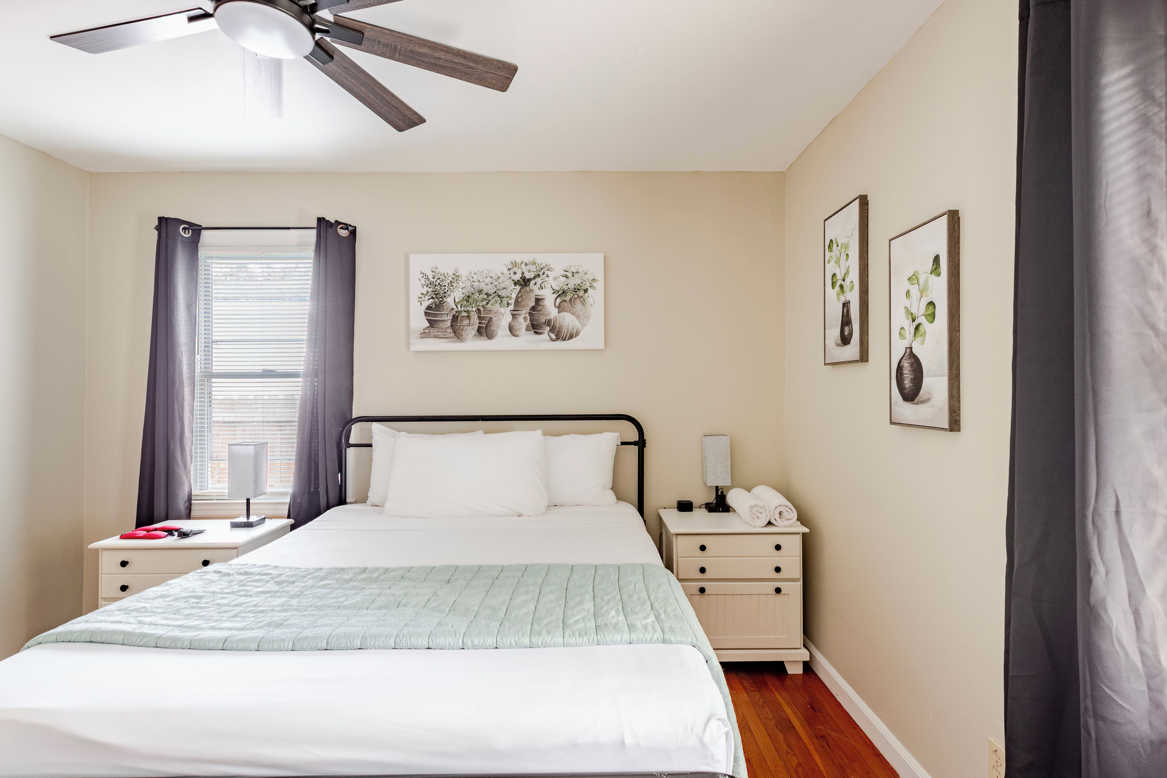 A bedroom with a bed, a nightstand, a bedside table, a ceiling fan, and a window with curtains.