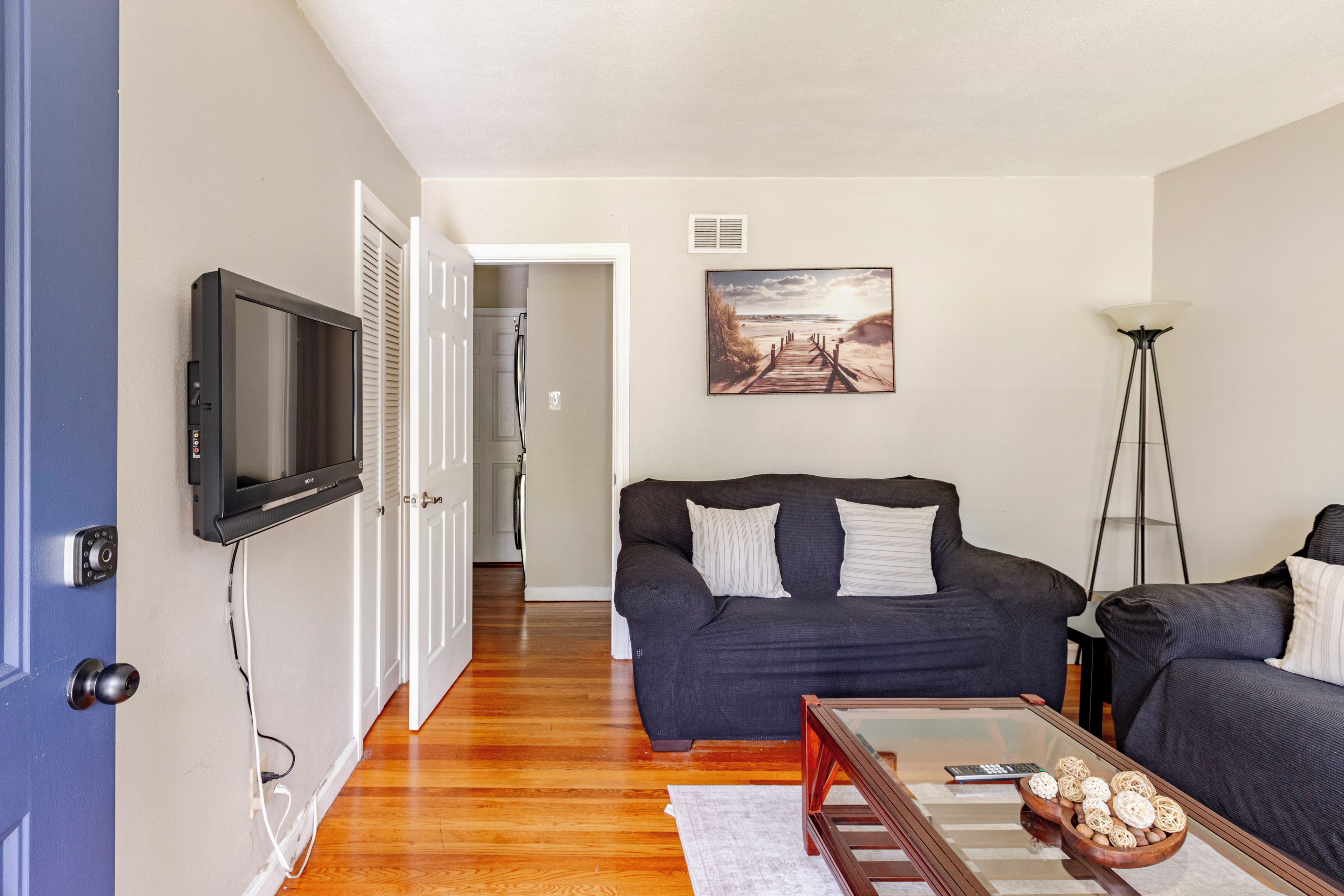 A living room with a dark-colored sofa, a flat-screen TV mounted on the wall, a wooden coffee table with decorative items, and a painting of a beach scene.