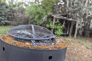 A portable grill with a mesh cover, surrounded by wood chips and debris, set in an outdoor area with trees and a wooden structure in the background. - Thunderbolt Coastal Cottage (Thunderbolt)