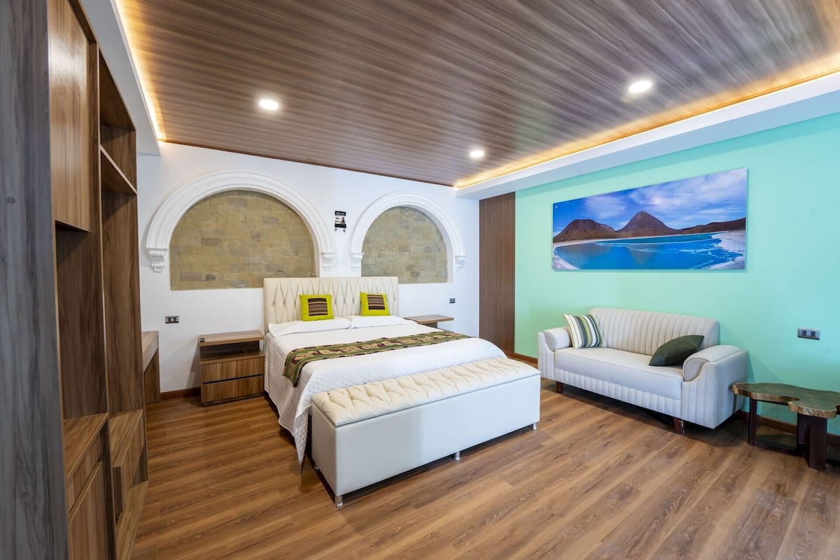 Un dormitorio con cama, sofá, suelo de madera, un cuadro y techo de madera.