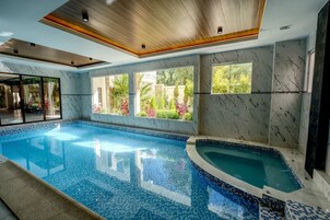 Una piscina con piso y paredes de baldosas, ventanas amplias con vista a follaje y techo de madera.