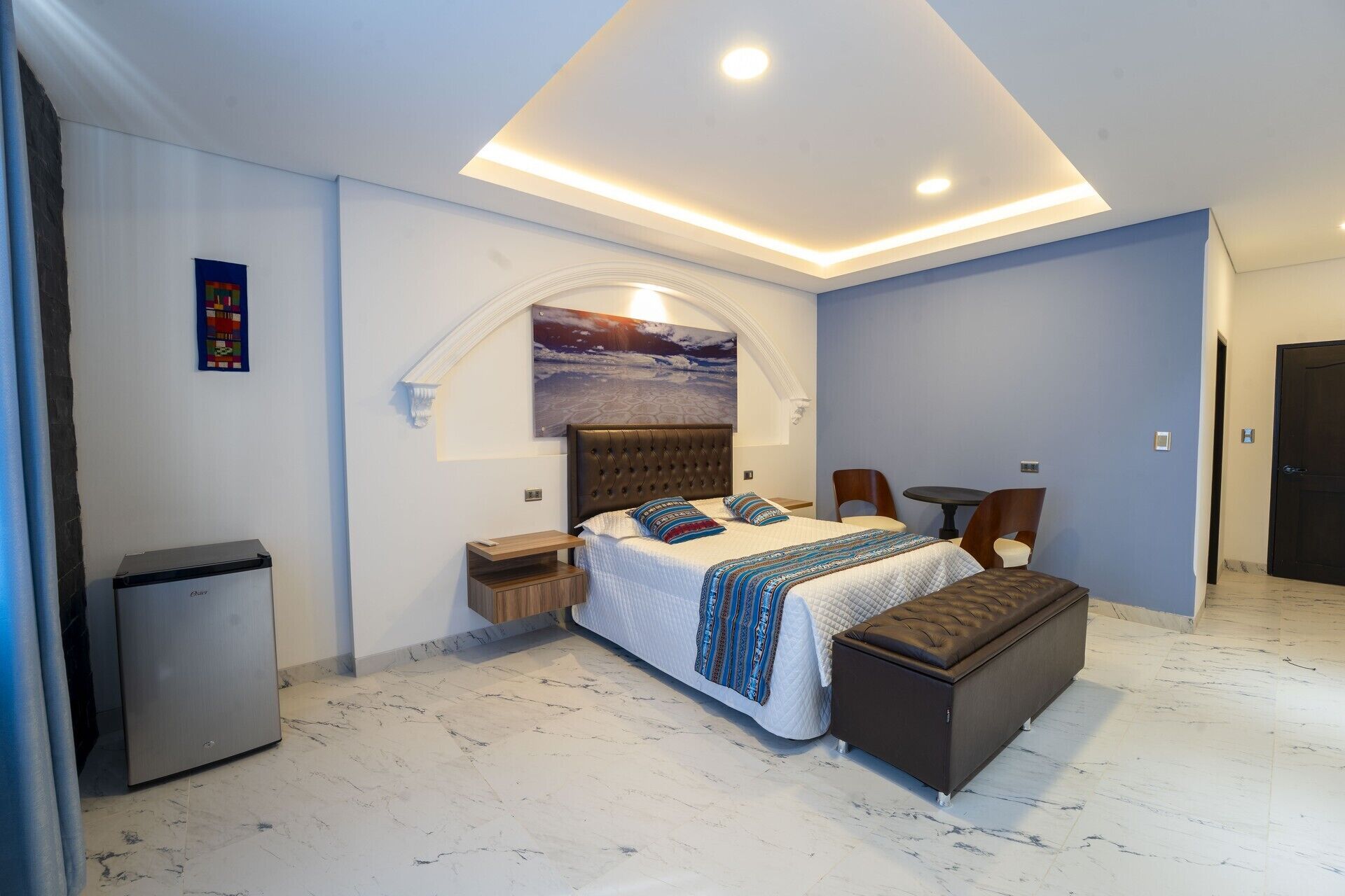 Een moderne slaapkamer met een bed, nachtkastjes, een bankje, een klein tafeltje en een deur.