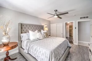 Ein Schlafzimmer mit einem gesteppten Kopfteil, einem Bett mit weißer Decke, einem Nachttisch mit einer Vase, einem Deckenventilator und einem begehbaren Kleiderschrank.
