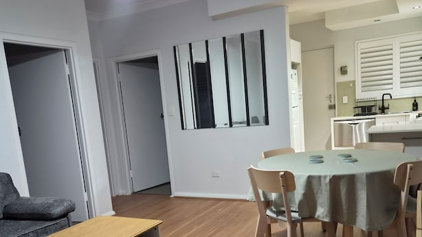 A modern living room with a dining area, wooden flooring, a ceiling fan, and a kitchen visible through an open door.