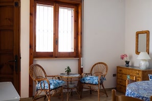 Ein Zimmer mit zwei Korbstühlen, einem kleinen Tisch, einem Schminktisch und einem Fenster mit Holzjalousien.