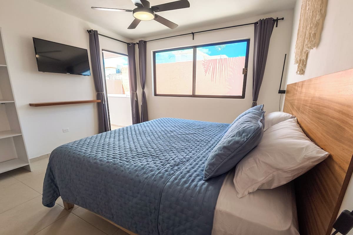 A bedroom with a bed, a ceiling fan, a TV, and a window with curtains.