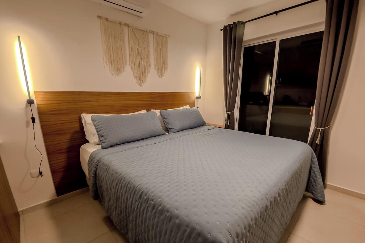 A neatly made bed with a headboard, two bedside lamps, and a window with curtains.