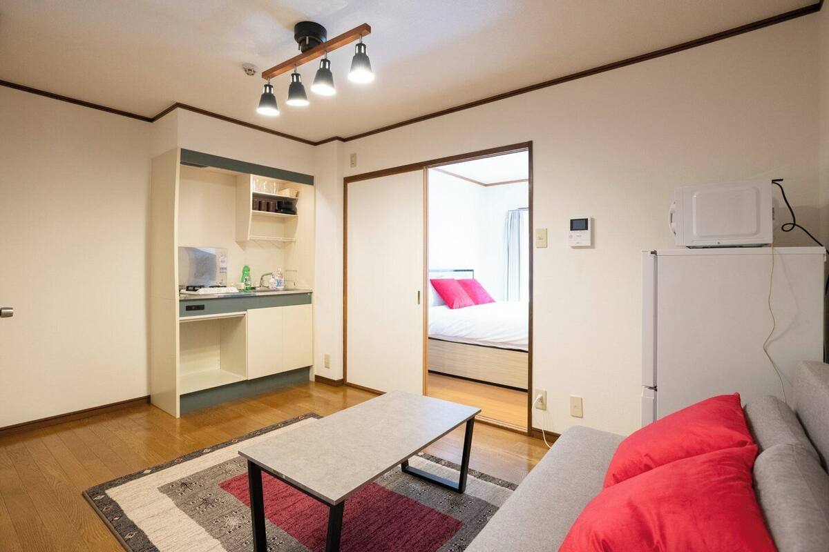 A compact hotel room with a kitchenette, a bed with red pillows, a small table, and a sofa.