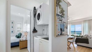 A room with a bed, a washing machine, a dining area, and a view of the ocean. - Beachside Stay - Coastal Comfort - Walk to Surf @ Alex Seaside Resort (Sunshine Coast)