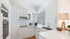 A modern kitchen with white cabinetry, a built-in oven, a sink, and a window with blinds. - Beachside Stay - Coastal Comfort - Walk to Surf @ Alex Seaside Resort (Sunshine Coast)