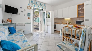 A compact living space with a sofa, dining table, chairs, a TV, kitchen area, and a door leading outside. - Baia Serena With Terrace & Parking (San Bartolomeo al Mare)