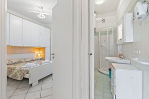 Habitación de hotel con cama, mesita de noche, ventilador de techo y baño con lavamanos, espejo y ducha.