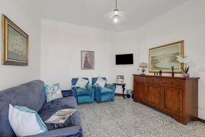 Een woonkamer met een bank, een blauwe fauteuil, een houten kast, een televisie en een ingelijst kunstwerk aan de muur.