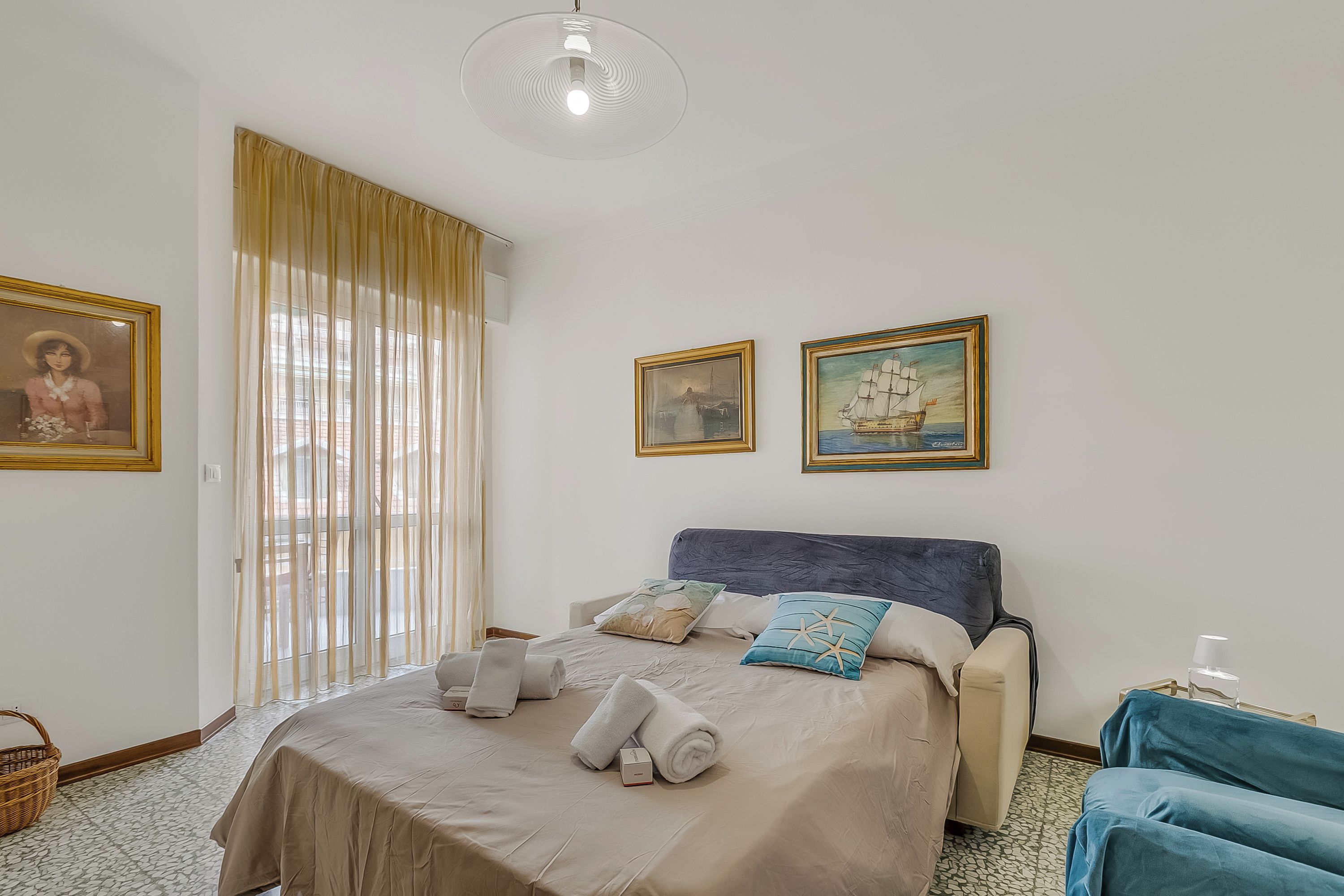 Ein ordentlich bezogenes Bett mit Handtüchern, ein blauer Stuhl und zwei gerahmte Bilder an der Wand.