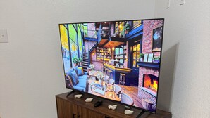 Ein Fernseher zeigt eine gemütliche Innenansicht, ein Holzschrank mit Fernbedienung, eine Aufbewahrungsbox mit dem Buchstaben 'S' und eine dekorative Vase mit Trockenpflanzen.