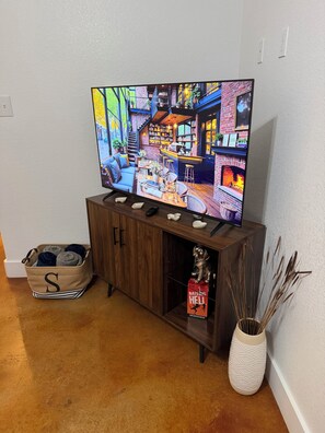 A television displaying a cozy interior scene, a wooden cabinet with a remote, a storage box with a letter 'S', and a decorative vase with dried plants. - StevensHill Waco • Baylor • Magnolia • Downtown • Backyard (Waco)