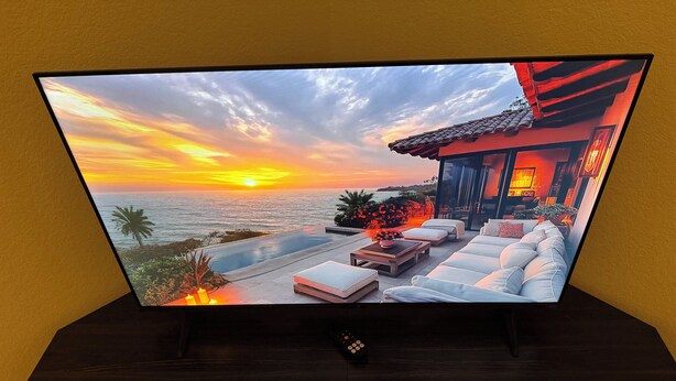 Ein Flachbildfernseher auf einem dunklen Holzständer zeigt einen Sonnenuntergang über dem Meer mit Sitzgelegenheiten im Freien.