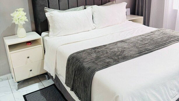 Una cama con cabecera oscura, ropa de cama blanca, una mesita de noche con un florero y una alfombra negra sobre suelo de baldosas.