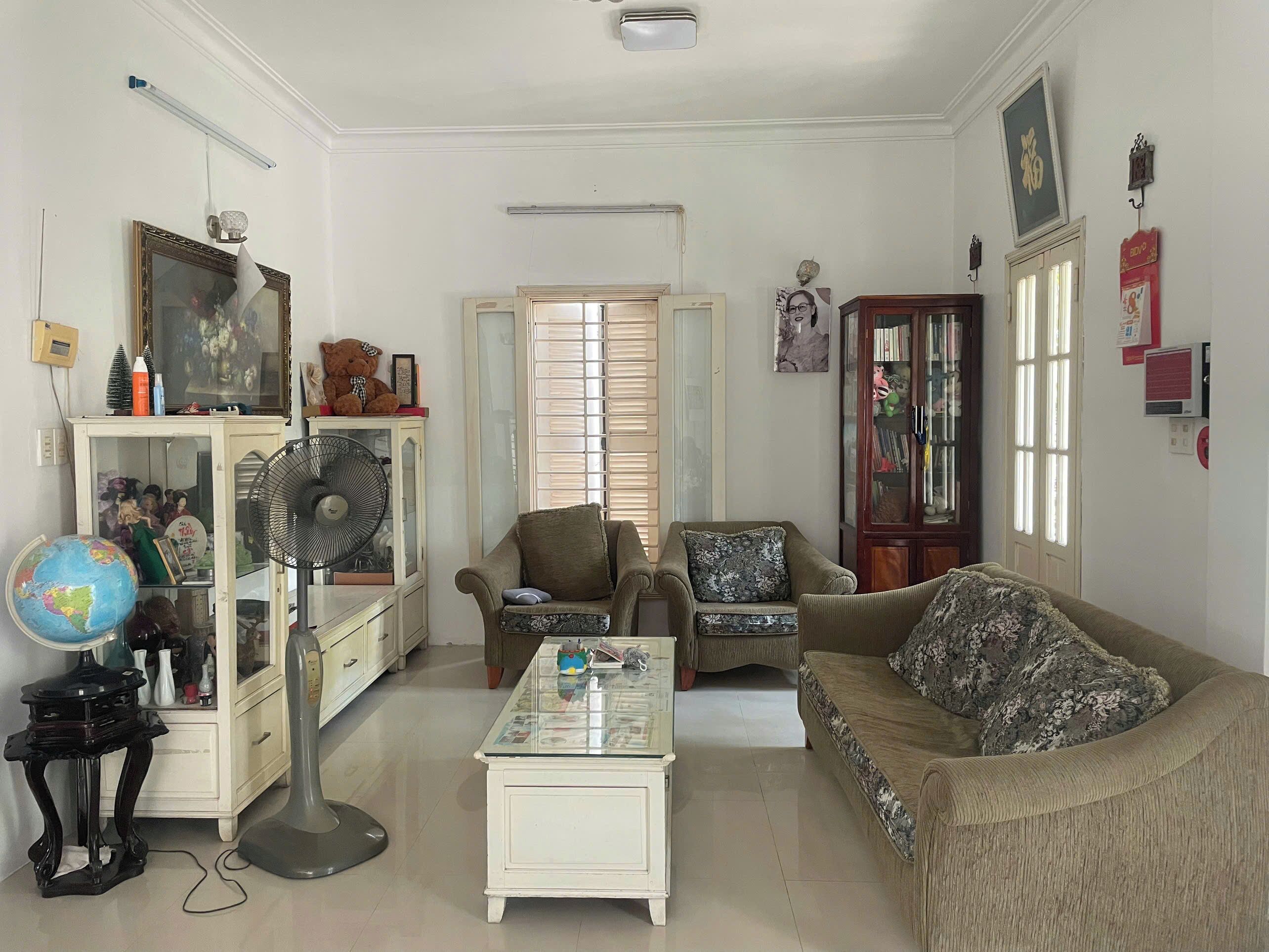 A living room with a sofa, armchair, a small table, a globe, a fan, and a cabinet with various items.