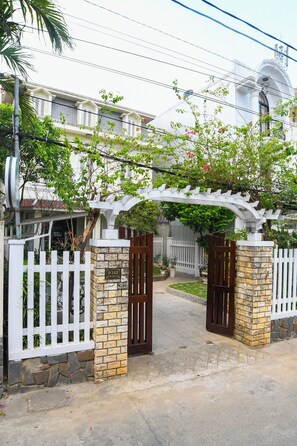 A gated entrance with a white picket fence, a stone pillar with a plaque, and a pergola with climbing plants. - Maison Vy Homestay (Phuong Vi Da)