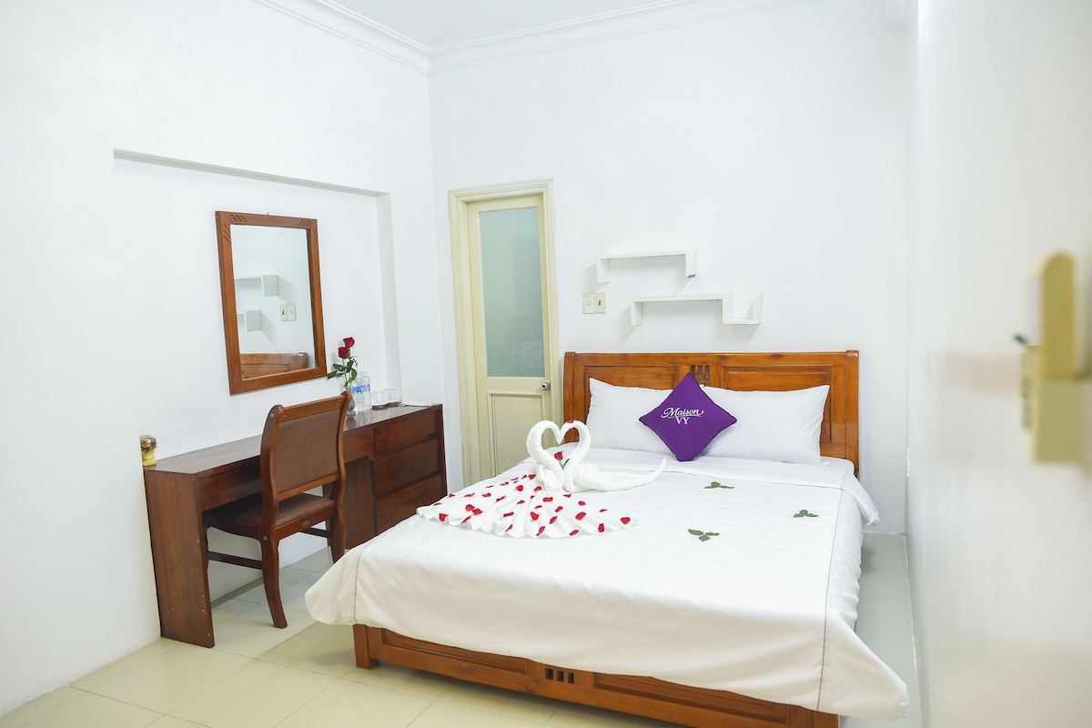 A hotel room with a bed, a wooden desk, a chair, a mirror, and a door.