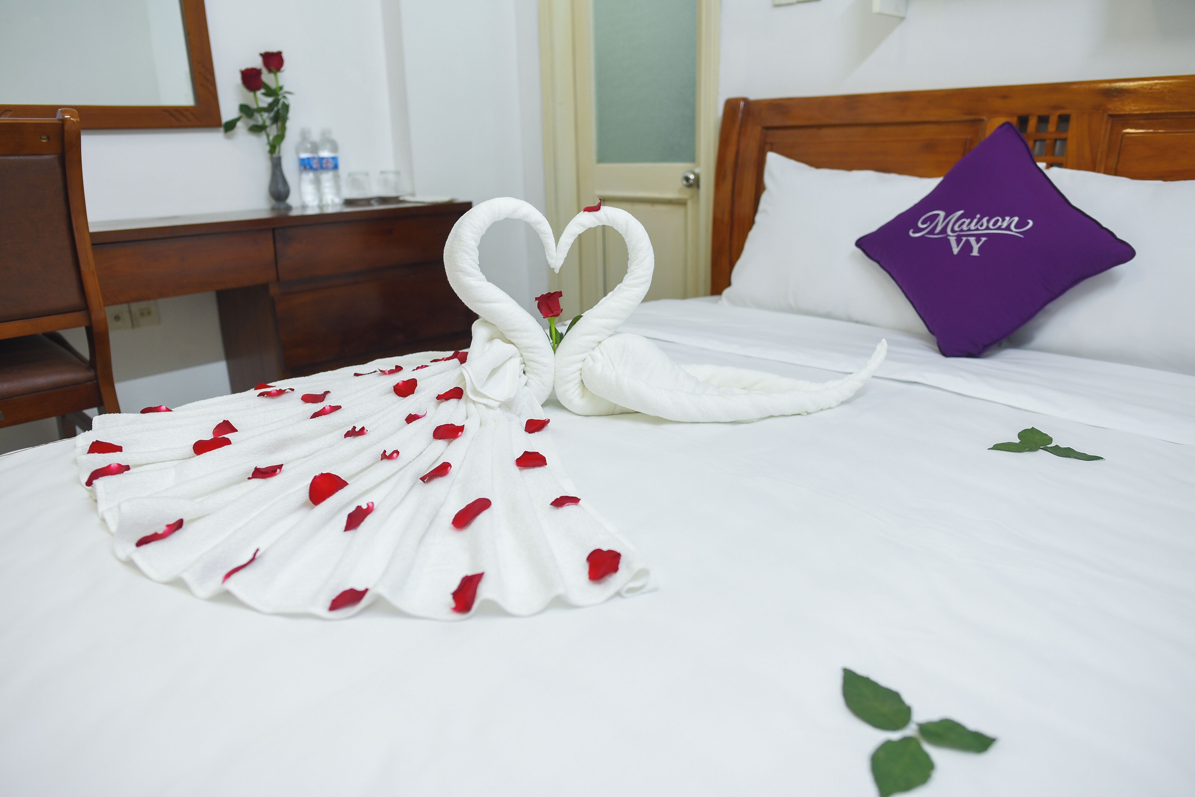 Uma cama com colcha branca, um travesseiro roxo com a inscrição 'Maison VY' e uma toalha arrumada em formato de cisne, decorada com pétalas de rosa vermelhas.