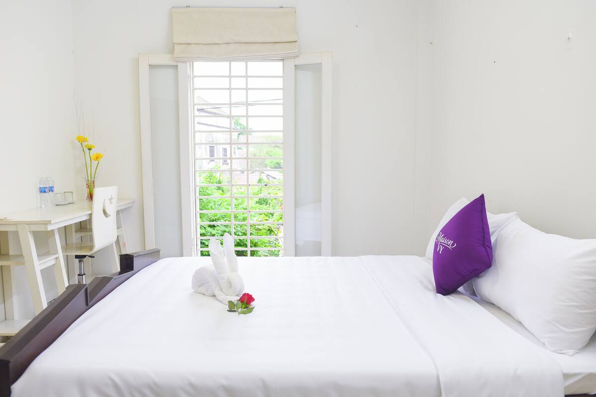 A hotel room with a bed, a desk, and a window with a view of greenery.