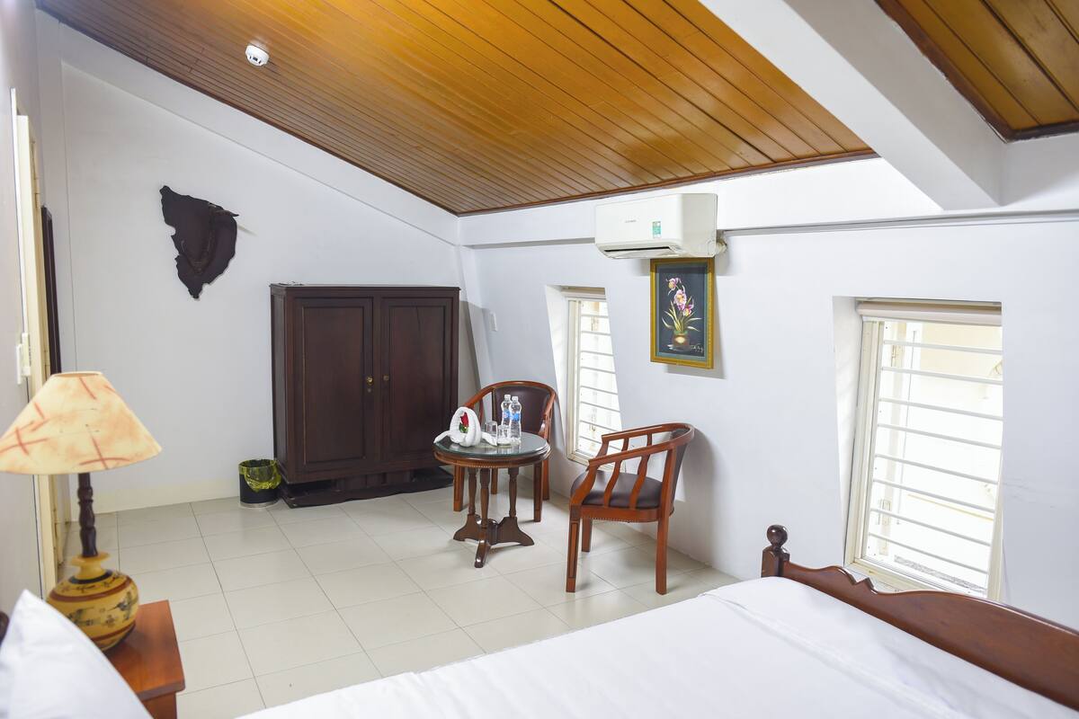 A bedroom with a bed, a wooden ceiling, a window, a chair, and a table with a lamp.