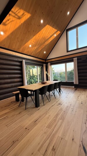 A spacious room with a wooden table and chairs, large windows, and a sloped ceiling.