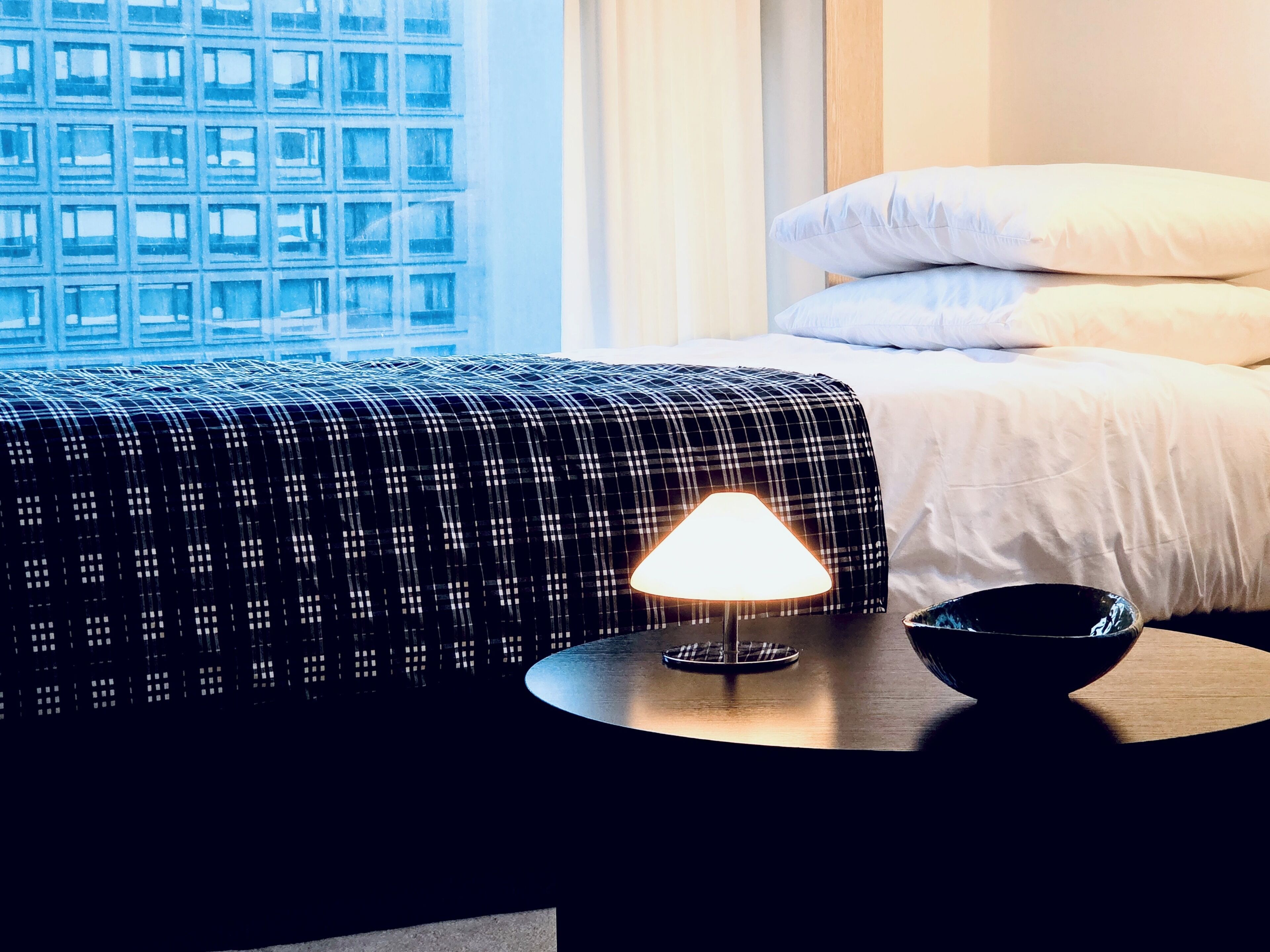 A hotel room with a bed, a bedside table with a lamp, a bowl, and a window with a view of a building.