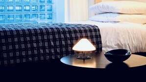 Ett hotellrum med en säng, ett nattduksbord med en lampa, en skål och ett fönster med utsikt över en byggnad.