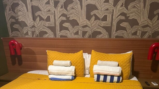 A hotel room with a bed covered in a yellow quilt, two pillows, and a neatly folded white towel. - 1818 LLC (San Juan)