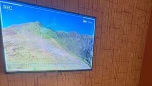 A flat-screen TV mounted on a brick wall, a wooden cabinet below it, and a red chair to the right. - 1818 LLC (San Juan)