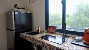 A compact kitchen with a refrigerator, a sink, and a window with a view of trees. - 1818 LLC (San Juan)